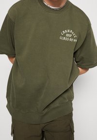 Sudadera verde oliva hecha de un tejido suave, con mangas cortas, cuello redondo y texto blanco "Carhartt MVP Clase del 89" en la parte frontal.