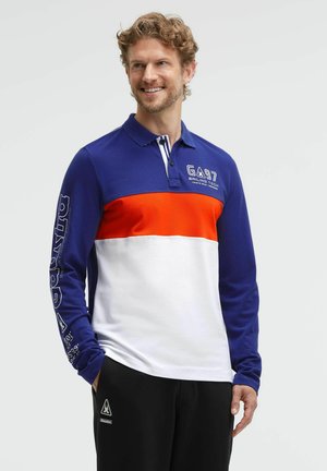 Smiling man with curly hair wearing a long-sleeve polo shirt with blue, orange, and white color blocks and black pants, hands in pockets.