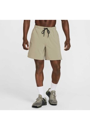Uomo che indossa pantaloncini beige con coulisse nera, calzini bianchi a coste e scarpe da ginnastica grigie, in piedi su sfondo bianco.