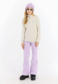 Lichtbeige quarter-zip top met geborduurd logo, gecombineerd met lichtpaarse broek en zwarte sneeuwlaarzen. Accessoires zijn een lavendelkleurige beanie en witte zonnebril.
