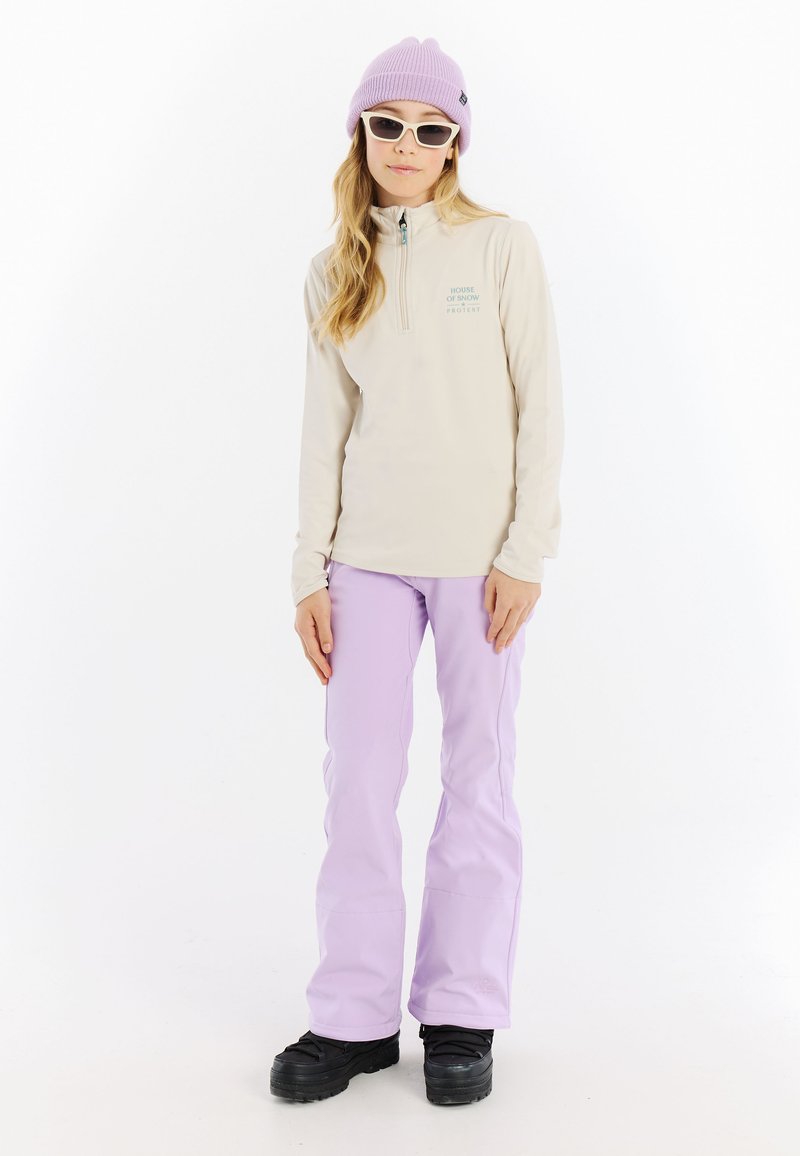 Lichtbeige quarter-zip top met geborduurd logo, gecombineerd met lichtpaarse broek en zwarte sneeuwlaarzen. Accessoires zijn een lavendelkleurige beanie en witte zonnebril.