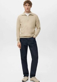 Beige trui met rits en omgeslagen kraag, gecombineerd met donkerblauwe spijkerbroek en beige sneakers. Model staand tegen een effen achtergrond.