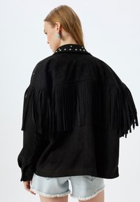 Zwarte faux-suède jas met franjes aan de achterkant en zilveren studs op de kraag. Gestyled met denimshorts.