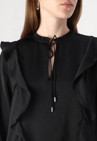 Blouse noire avec manches à volants, encolure froncée avec un détail à nouer, et un léger éclat. Confectionnée dans un tissu léger.