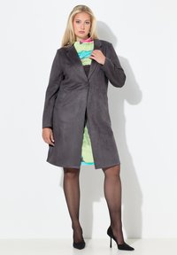 Abrigo de ante gris con cierre de un solo botón y bolsillos laterales, combinado con un vestido estampado multicolor y tacones altos negros.