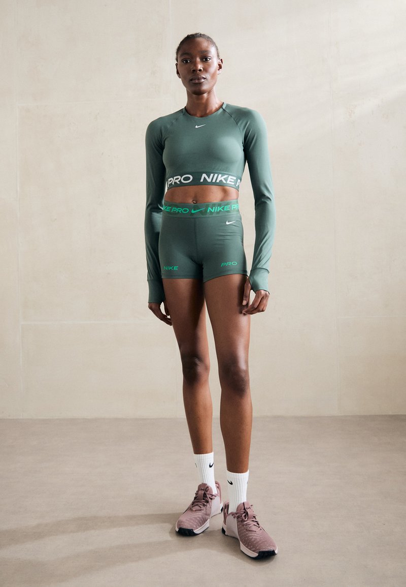 Top corto verde a maniche lunghe e pantaloni corti a vita alta abbinati con loghi Nike neri e verdi. Il modello indossa sneaker rosa chiaro e calze bianche.