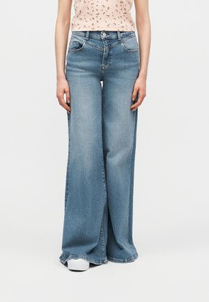 Γυναίκα που φοράει φαρδιά μπλε τζιν παντελόνια και λευκά αθλητικά παπούτσια, στέκεται μπροστά σε ένα απλό ανοιχτό φόντο.