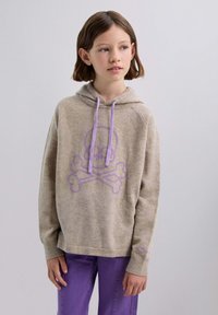 Beige Kapuzenpullover mit einem lila Schädelsymbol und gekreuzten Knochen, lila Kordeln und einem lockeren Schnitt. Der Stoff wirkt weich und strukturiert.