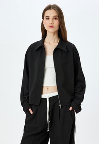 Giacca nera con zip e colletto, realizzata in tessuto testurizzato. Abbinata a un top bianco a coste e pantaloni neri con coulisse.