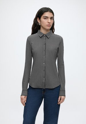 Γυναίκα με μια μαύρη και λευκή κάθετα ριγέ πουκάμισο με κουμπιά και μακριά μανίκια και μπλε τζιν, στέκεται μπροστά σε λευκό φόντο.