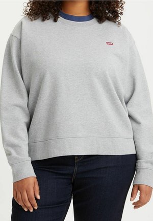Osoba s kudrnatými vlasy, která má na sobě světle šedou mikinu Levi's s námořnicky modrým límcem a tmavě modré džíny, stojící před jednobarevným pozadím.