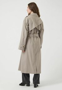 Trench coat largo de color beige, hecho de tejido, con una cintura ceñida por un cinturón, grandes solapas y puños abotonados; textura suave con un caído fluido.
