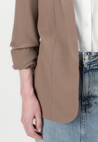 Brazo izquierdo de una persona que lleva una chaqueta marrón arremangada, una camisa blanca y unos vaqueros de mezclilla azules con detalles de botones y bolsillos visibles.