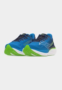 Μπλε και πράσινα παπούτσια τρεξίματος με μα ύρα κορδόνια, λευκές μεσαίες σόλες και λογότυπο της μάρκας στα πλάγια, εμφανιζόμενα από χαμηλή μπροστινή γωνία σε λευκό φόντο.