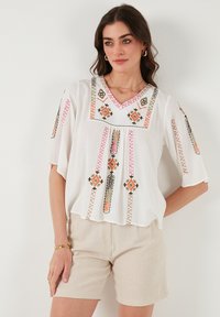 Blusa blanca de manga corta con intrincados bordados en rosa, naranja y verde, que presenta un escote en V y suaves pliegues en la parte frontal.