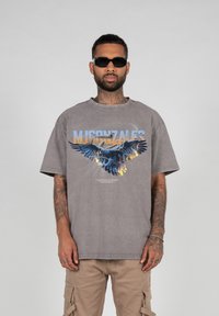 Grå oversized t-skjorte med et grafisk motiv av en ørn og ordet "MIGONZALES" i blått. Har rund hals og korte ermer.