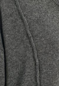 Maglione grigio lavorato a maglia con una consistenza morbida, bordi a coste e un evidente dettaglio di cucitura lungo il lato. Il tessuto è un mix caldo e flessibile.