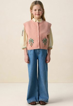 Jeune fille portant un gilet en maille rose sans manches avec une broderie de palmier, une blouse crème à manches bouffantes, un large jean bleu et des chaussures marron.