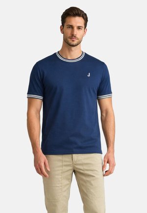 Uomo che indossa una t-shirt blu navy a maniche corte con colletto e polsini a righe bianche, abbinata a pantaloni beige, in piedi davanti a uno sfondo chiaro.