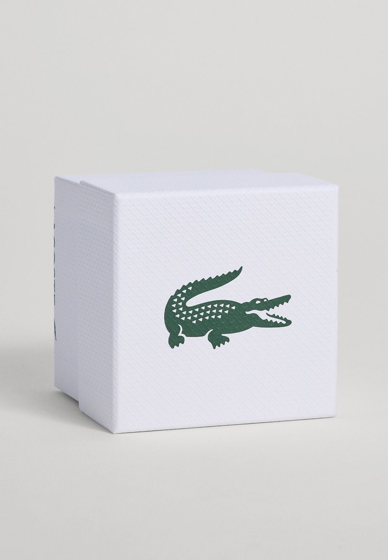 Weiße strukturierte Box mit grünem Krokodil-Logo, zentriert auf der Vorderseite vor einem einfarbigen hellen Hintergrund.