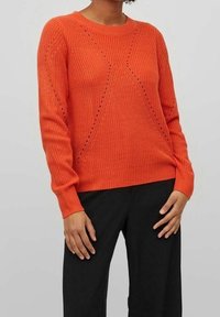 Oranger Strickpullover mit rundem Ausschnitt, strukturiertem Muster mit diagonalen Linien und langen Ärmeln, kombiniert mit schwarzer Hose.