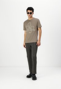 Brown graphic t-shirt with white print, paired with textured dark gray trousers. Model wears black sunglasses and black shoes. Minimalist setting.