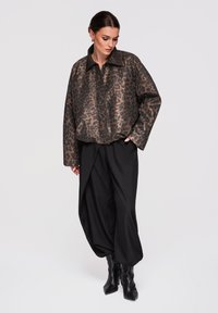 Femme posant en veste marron à imprimé léopard, pantalon noir ample et bottes noires pointues sur fond uni.