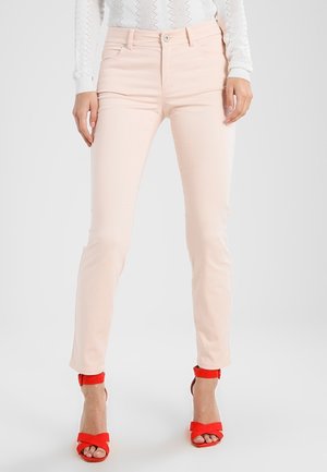 Mujer con pantalones ajustados rosa claro y tacones rojos abiertos, con una blusa blanca de manga larga y textura, de pie frente a un fondo gris.