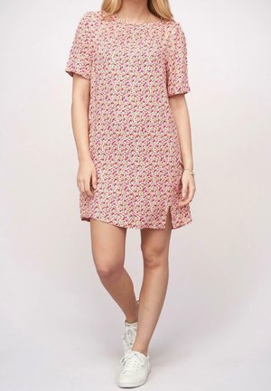 Nainen kävelee lyhythihainen, vaaleanpunainen kukallinen mekko ja valkoiset tennarit yhdistettynä, yksinkertaista vaaleaa taustaa vasten.