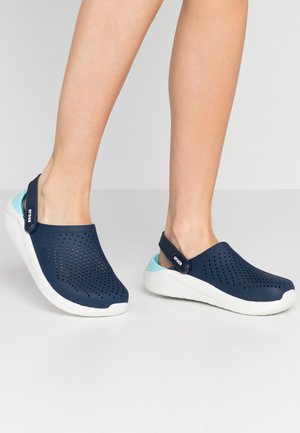 Beine in marineblauen Slip-on-Clogs mit perforierten Obermaterialien, weißen Sohlen und verstellbaren Fersenriemen, stehend auf einem hellgrauen Boden.