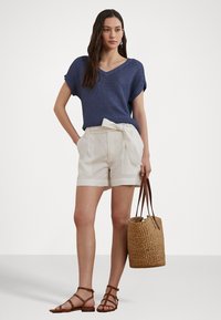 Top de punto azul con escote en V, pantalones cortos de lino blancos con lazo en la cintura, sandalias marrones de tiras y un bolso de mimbre. Modelo de pie contra un fondo liso.