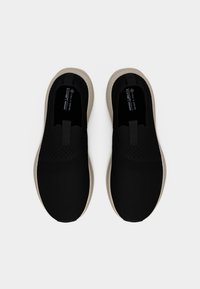 Μαύρα slip-on αθλητικά παπούτσια με λευκές σόλες και αεριζόμενο πλεκτό ύφασμα, που φαίνονται από πάνω, με ετικέτα vegan και άνεση αφρώδους πάτους.