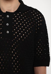 Polo noir en tricot avec motif ajouré, doté d'un col et de trois boutons circulaires, manches courtes et apparence texturée.