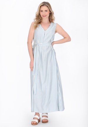 Vrouw met blond haar, die een mouwloze maxi-jurk met blauwe en witte verticale strepen draagt en witte platformsandalen aanheeft, poserend met haar hand op haar heup.