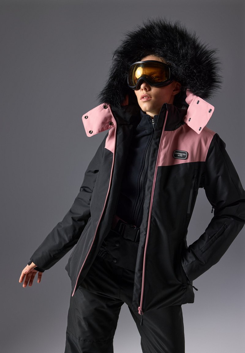 Svart og rosa skijakke med hette med pelskant, glidelås foran og trykknapper. Har flere lommer og kontrasterende glidelåsdetaljer.