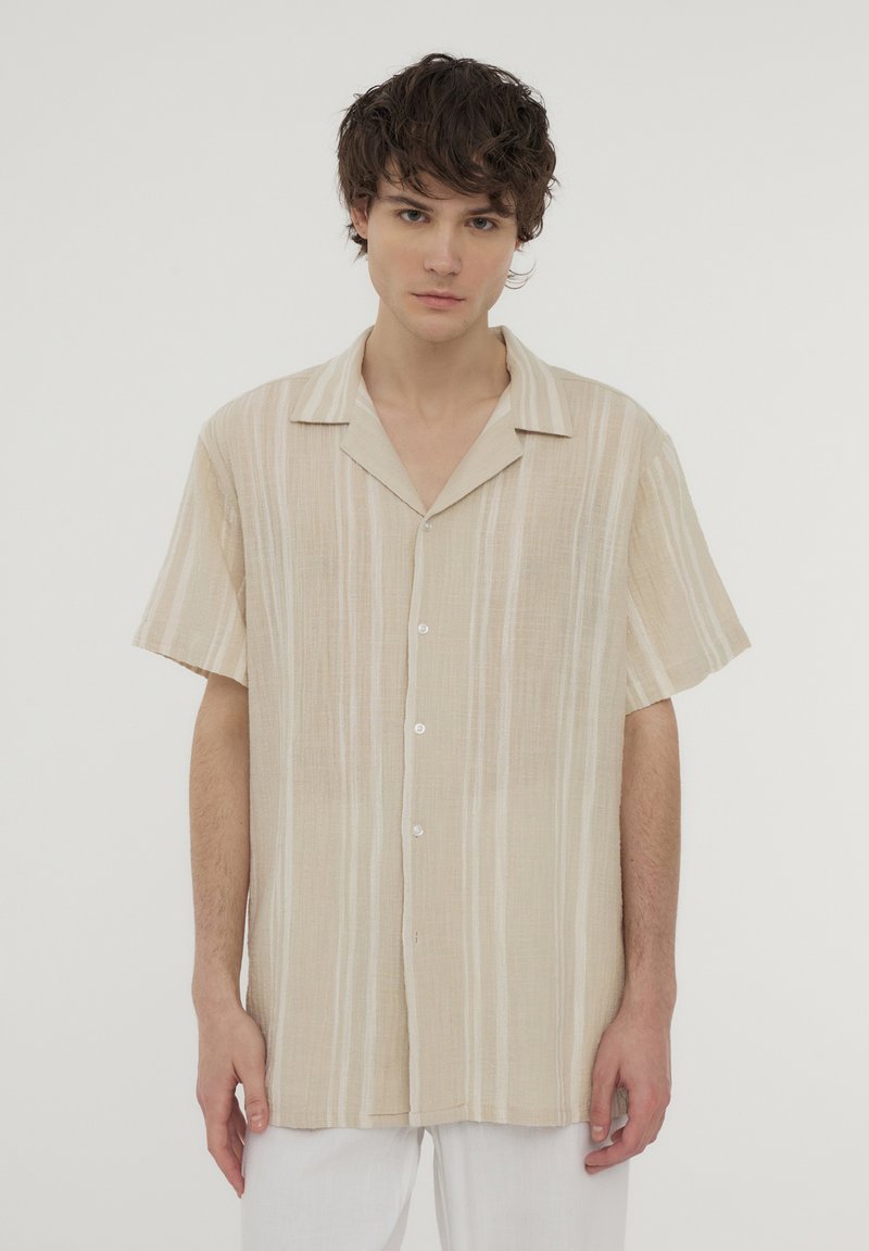 Camicia beige a maniche corte con strisce bianche verticali, tessuto leggero e chiusura frontale con bottoni. Presenta un classico design con colletto.