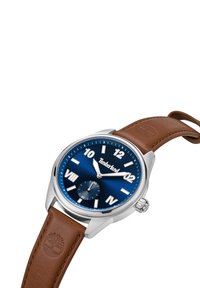Reloj de acero inoxidable plateado con una esfera azul oscura, números árabes blancos y una correa de cuero marrón con un logo grabado.