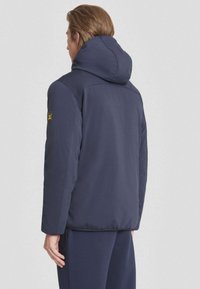 Veste à capuche bleu marine avec une texture lisse, des poignets élastiques et un détail de logo subtil sur la manche gauche. Conçue pour une silhouette ajustée.