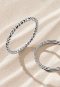 Zwei silberne Ringe auf einer hellen Stoffoberfläche, einer gedreht und der andere glatt, werfen sanfte Schatten.