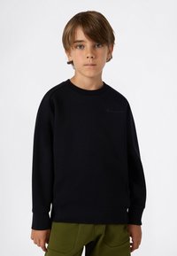 Ragazzo con capelli castano chiaro che indossa un felpa nera e pantaloni verde oliva, in piedi davanti a uno sfondo bianco semplice.