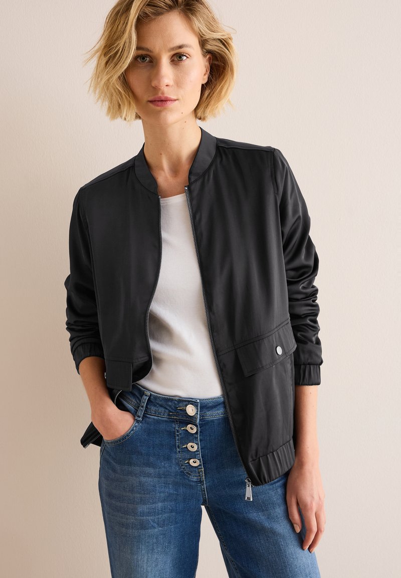 Veste bomber noire avec un col rond, fermeture éclair à l'avant, poignets élastiques et une poche sur le côté. Associée à un jean bleu avec des détails de boutons.