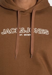 Brown cotton hoodie featuring white text "JACK&JONES" and "EST. MCMXC" with matching drawstrings and metal tips. Textured fabric.