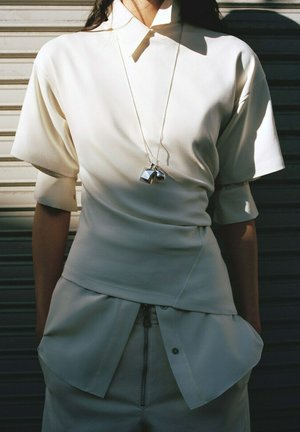 Person trägt übereinander geschichtete weiße Kurzarmoberteile, hochtaillierte Hosen mit Reißverschluss und eine silberne Anhängerkette vor einem metallischen Hintergrund.