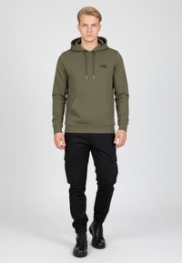 Giovane uomo che indossa un hoodie verde oliva con tasca frontale e pantaloni cargo neri, che cammina in avanti su uno sfondo chiaro e uniforme.