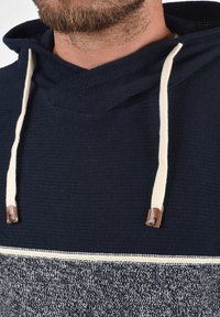 Sweat à capuche navy avec une texture tricotée, comprenant des cordons de serrage crème et des accents en cuir marron. La partie inférieure présente un motif texturé plus clair.