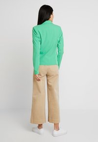 Veste ajustée verte avec un col, associée à un pantalon beige ample. La tenue est complétée par des baskets blanches. Tissu lisse et contemporain.