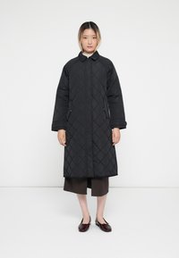 Black quilted long coat with a high collar, featuring a straight cut, large sleeves, and zippered pockets. Worn over brown trousers and flats.