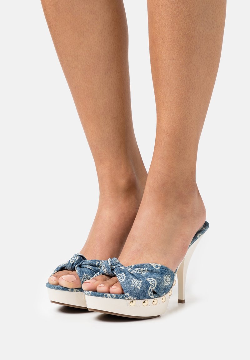 Pieds portant des talons hauts en denim bleu à bout ouvert avec un détail nœud et des semelles compensées blanches décorées de clous dorés.