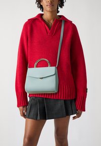 Borsa a mano in pelle azzurro chiaro con manico circolare, forma rettangolare, chiusura a patta e accessori dorati, indossata con un maglione rosso a maglia e una gonna a quadri.