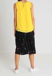 Gul ærmeløs top med en rygdetalje i form af et nøglehul, parret med en sort nederdel med farverige blomsterprint. Hvide sneakers fuldender looket.